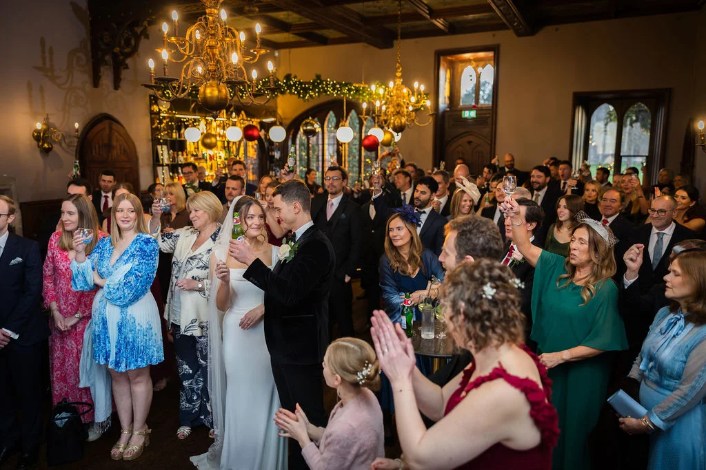 Newlyweds stand amidst a joyful crowd of wedding guests holding drinks and cheering inside a grand historic reception room featuring ornate chandeliers.