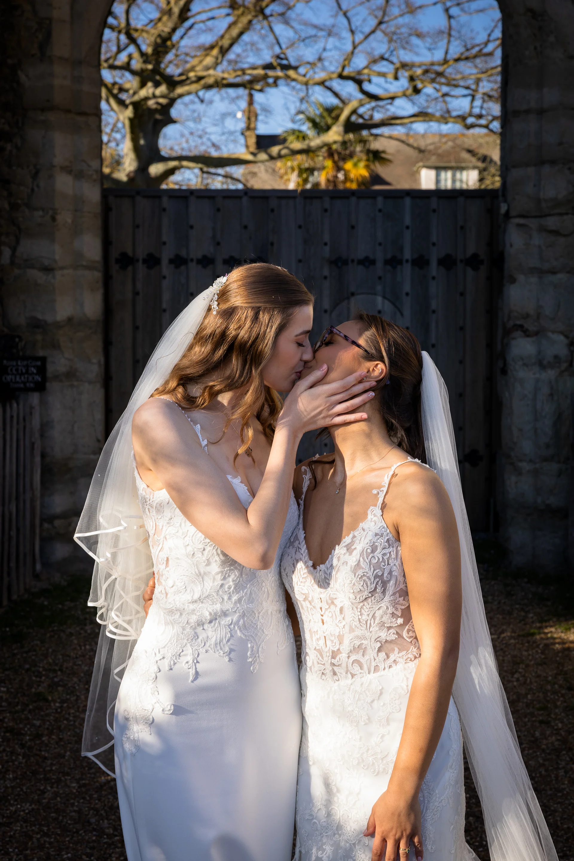Two brides share a tender kiss in front of a historic stone archway and wooden gates on a bright sunny wedding day.