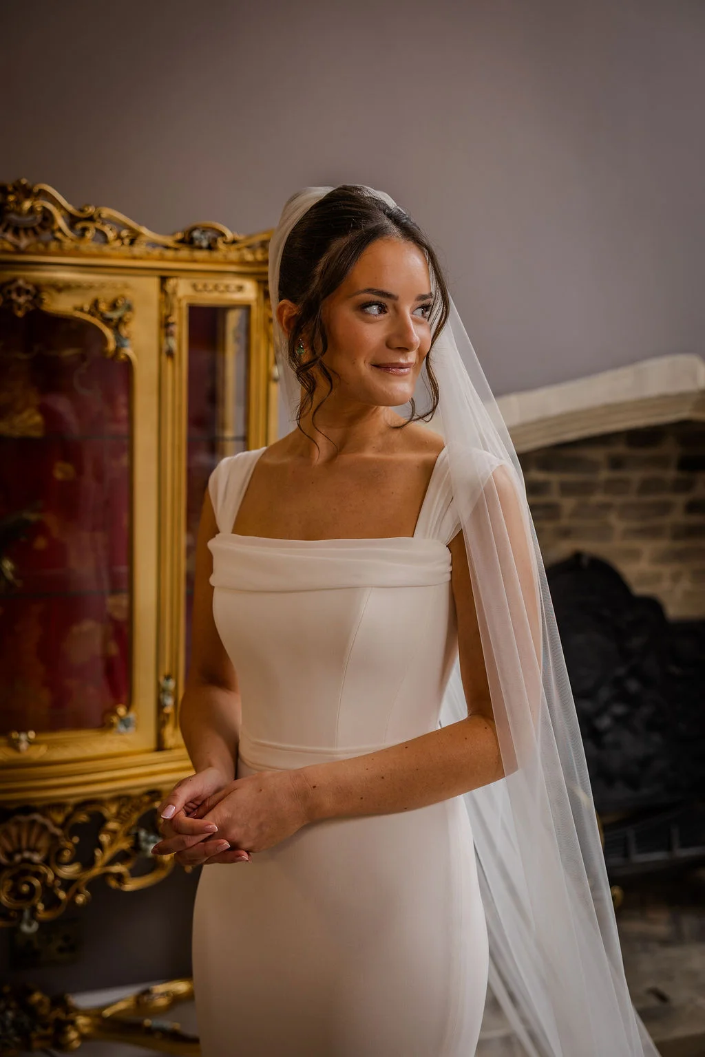A stunning bridal portrait of a bride wearing a structured square-neck crepe gown and a long veil, looking away with a gentle smile indoors.