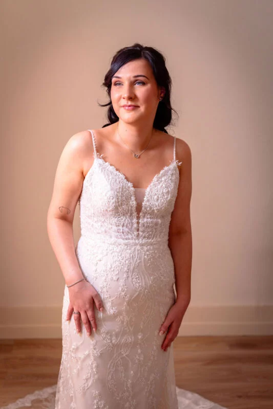 A graceful bride wearing a beaded lace a-line gown with spaghetti straps and a v-neckline poses indoors against a neutral wall.