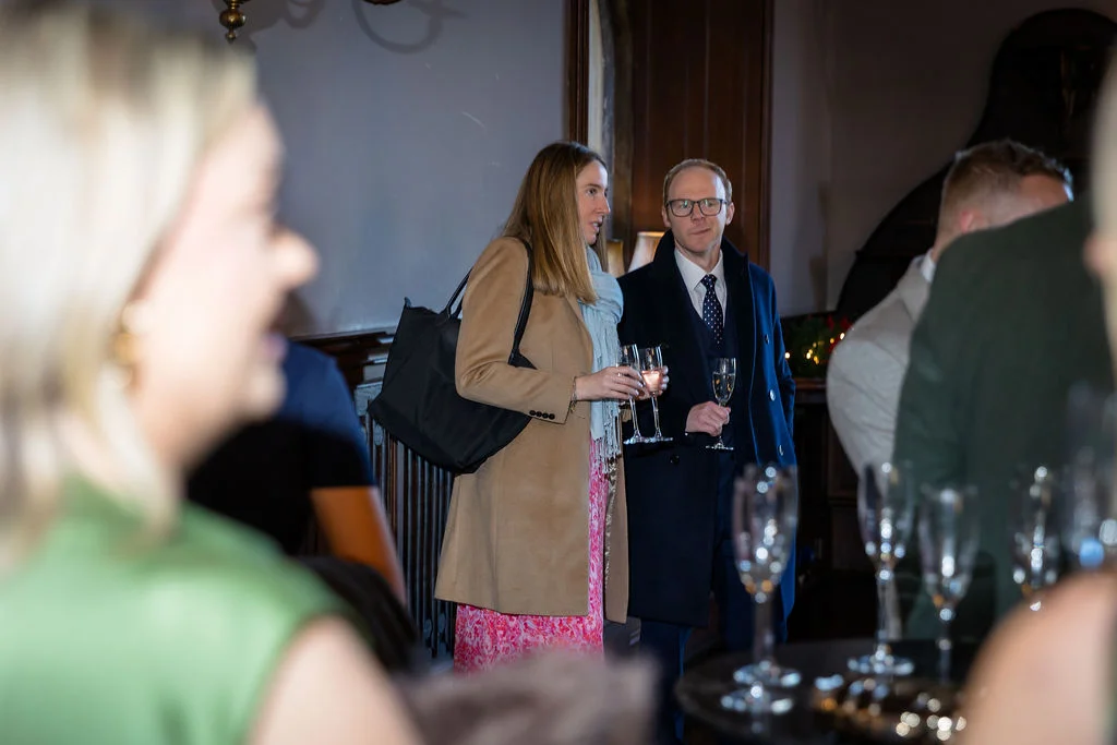 Wedding guests mingle indoors during a drinks reception, holding glasses of champagne while chatting in a warmly lit room.
