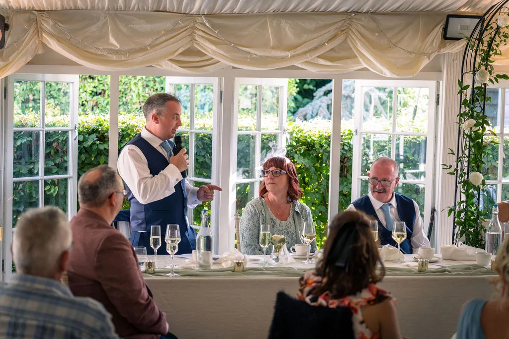A wedding guest stands to deliver a speech at the top table inside a brightly lit marquee draped with white fabric.