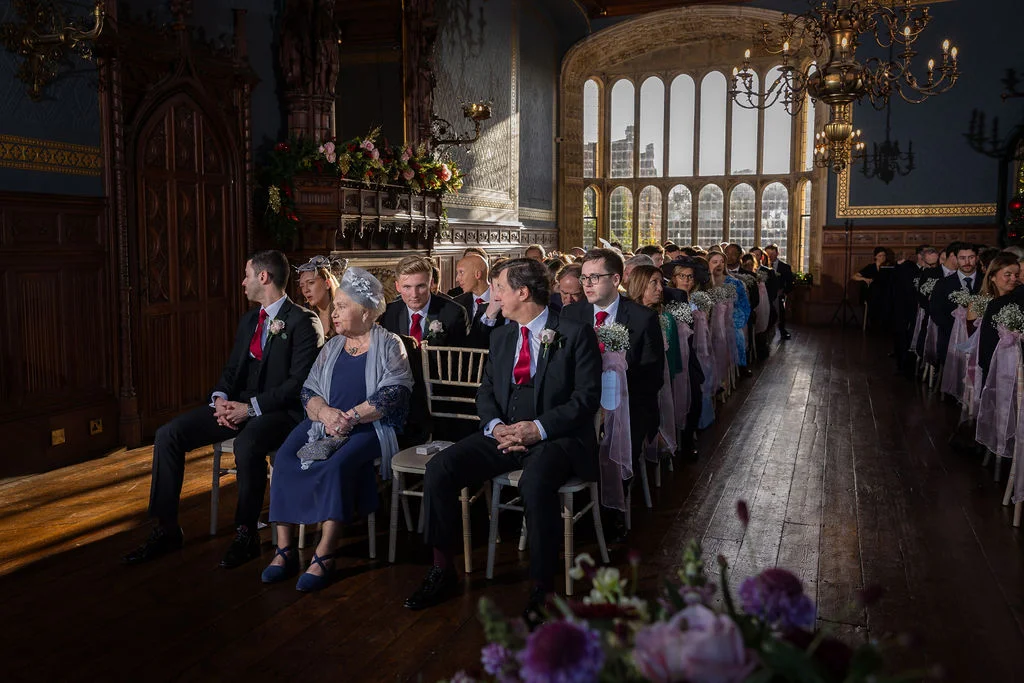 Seated wedding guests wait attentively inside a grand, wood-panelled hall illuminated by dramatic light streaming through tall, arched windows.