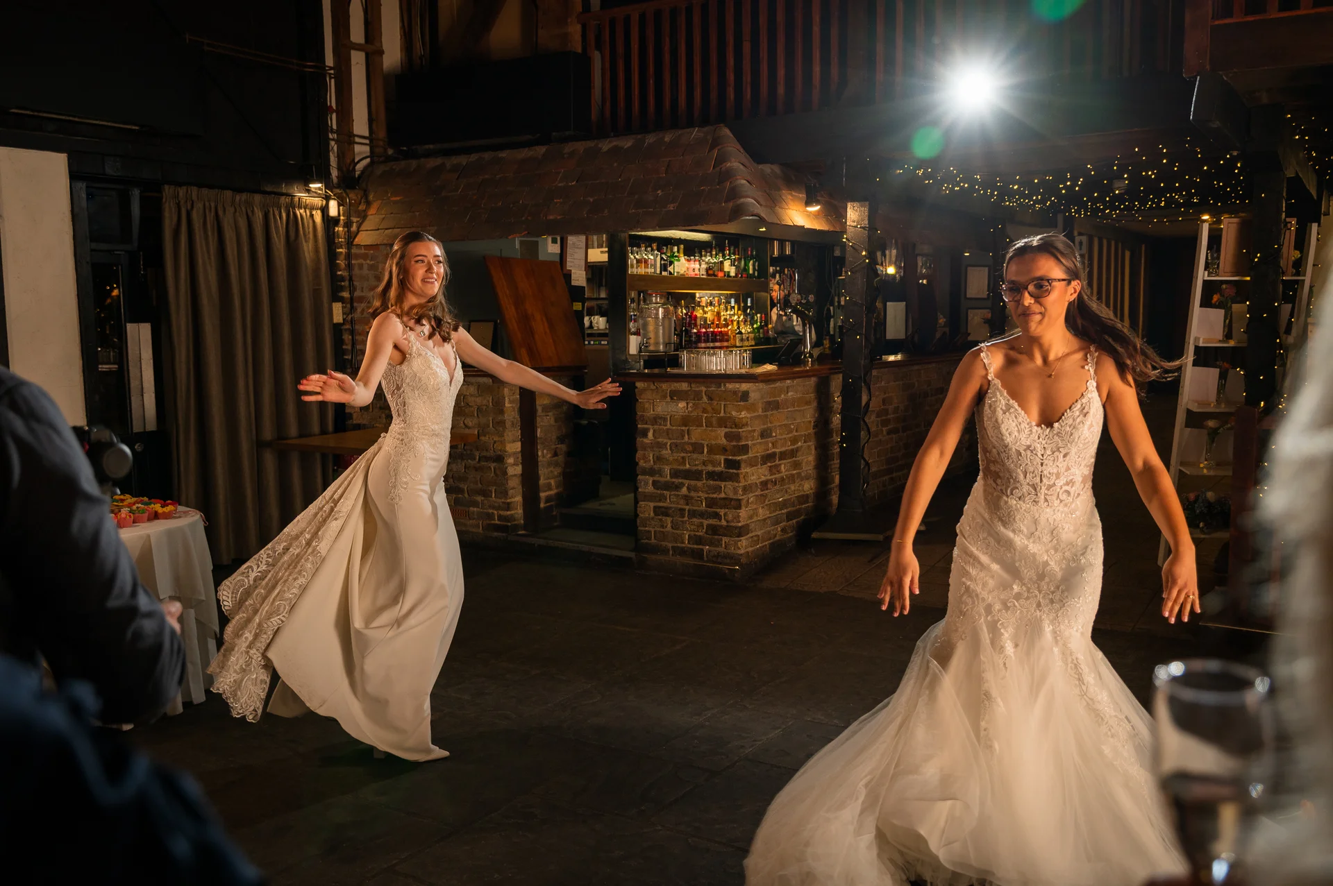 Two smiling brides in wedding dresses dance joyfully together inside a rustic barn reception venue illuminated by spotlights.