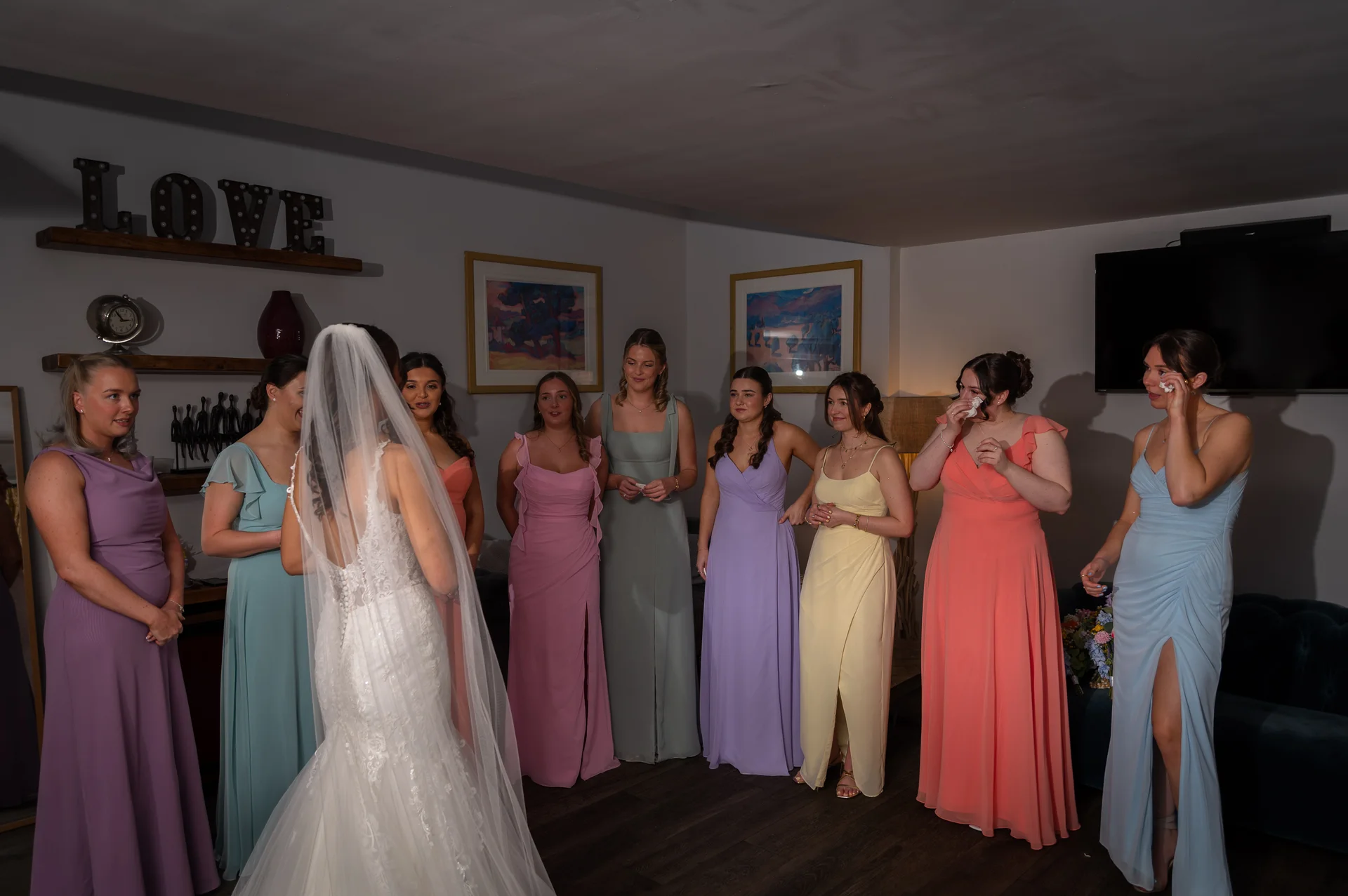 A stunning bride in a lace wedding dress with a long veil shares an emotional first look with her nine bridesmaids wearing colourful pastel dresses in a cosy indoor setting.