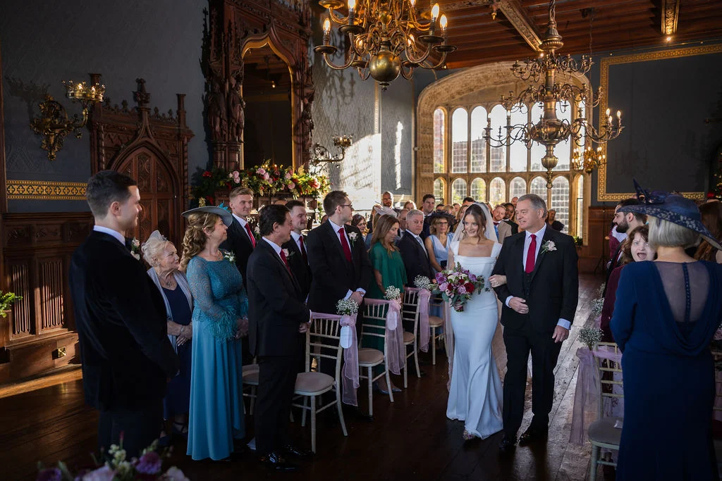 A bride walks down the aisle on her father's arm in a grand wood-panelled hall filled with seated guests and groomsmen.