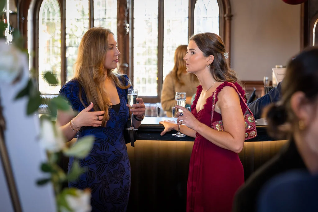 Two wedding guests in stylish dresses hold champagne flutes and chat near a bar during the drinks reception.