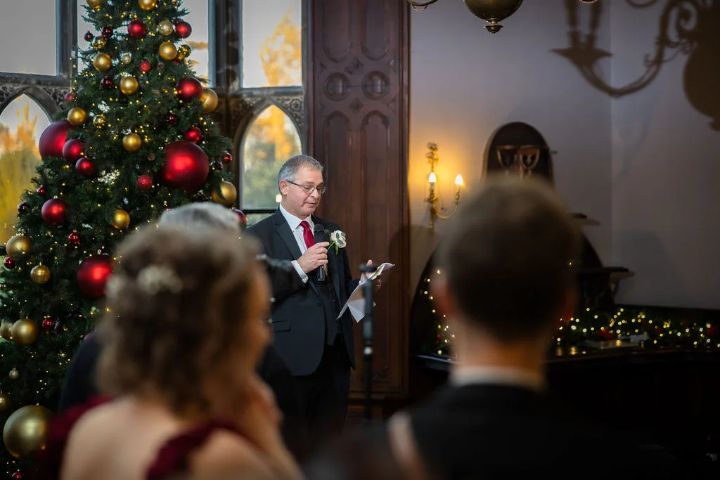 A father of the bride delivers his speech next to a large, festively decorated Christmas tree inside an elegant panelled room during a winter wedding reception.