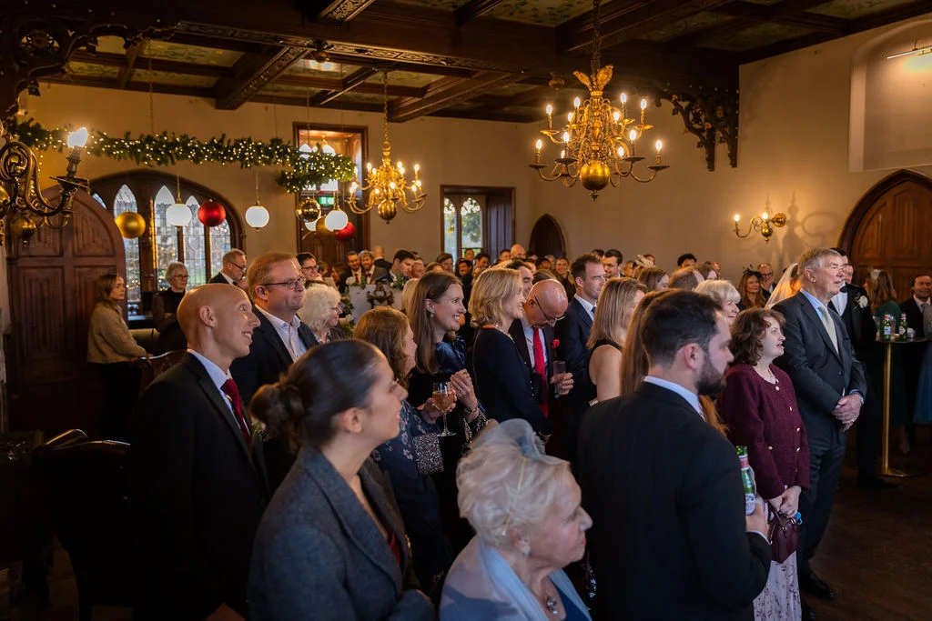 A packed room of wedding guests in a grand historic ballroom with ornate chandeliers and festive decorations watching attentively during the reception.