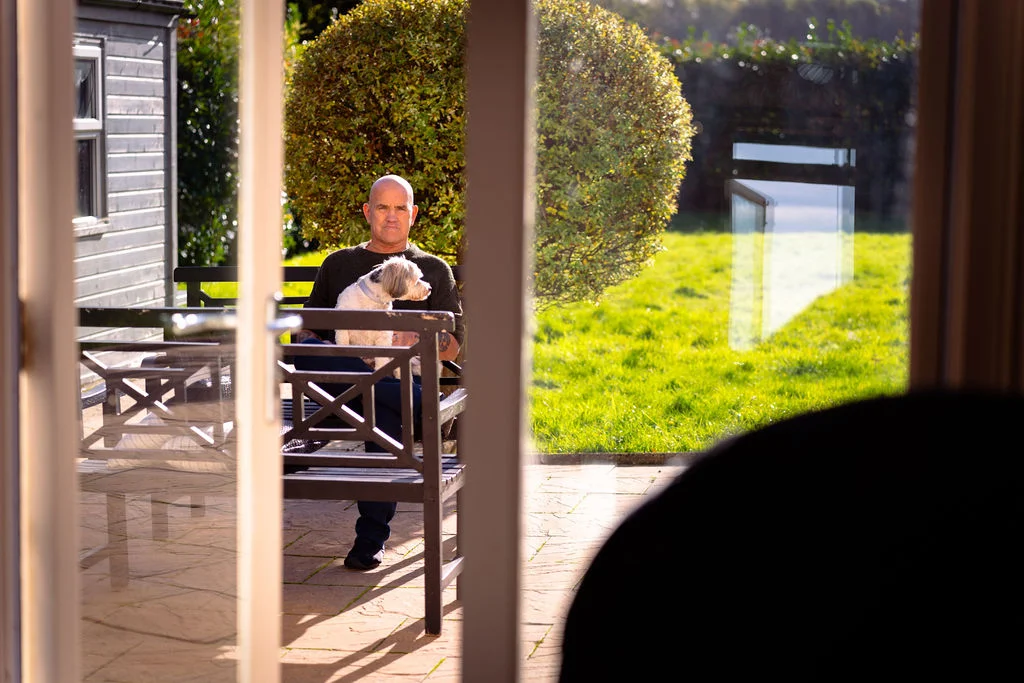 A man sits outside on a patio bench holding a small dog, viewed through a glass doorway from inside a building.