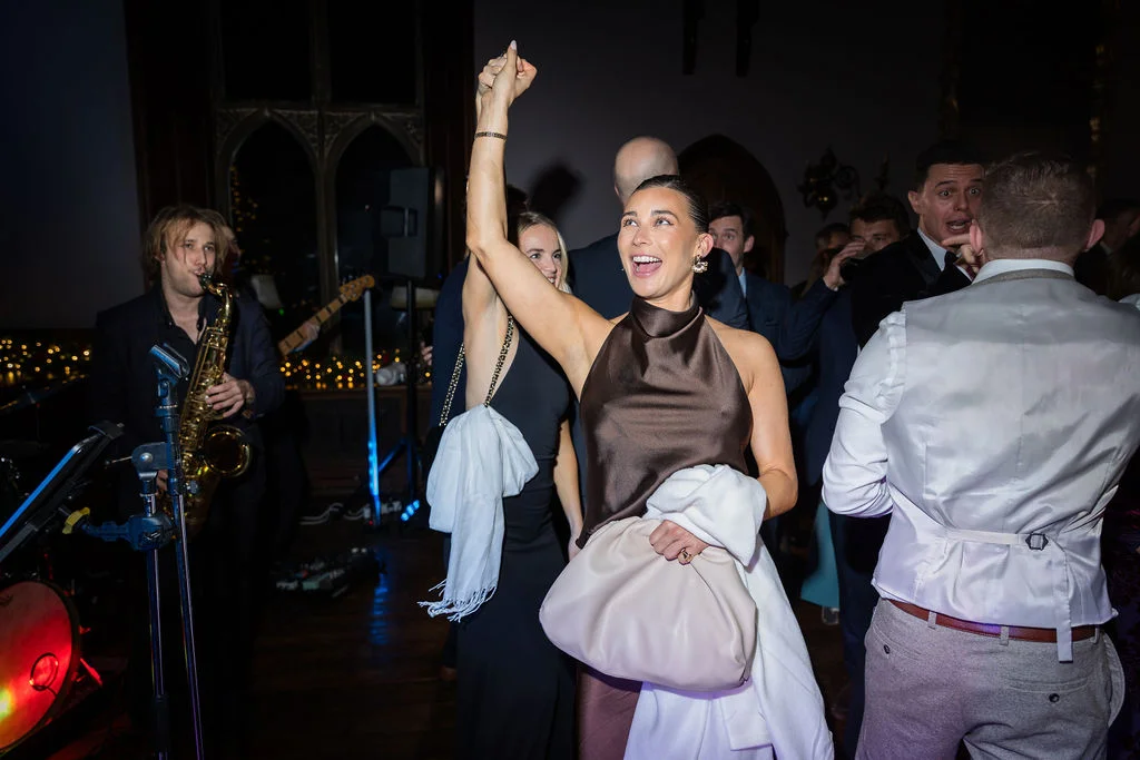 A joyful wedding guest with her arm raised in celebration on a lively dance floor as a saxophonist plays in the background.