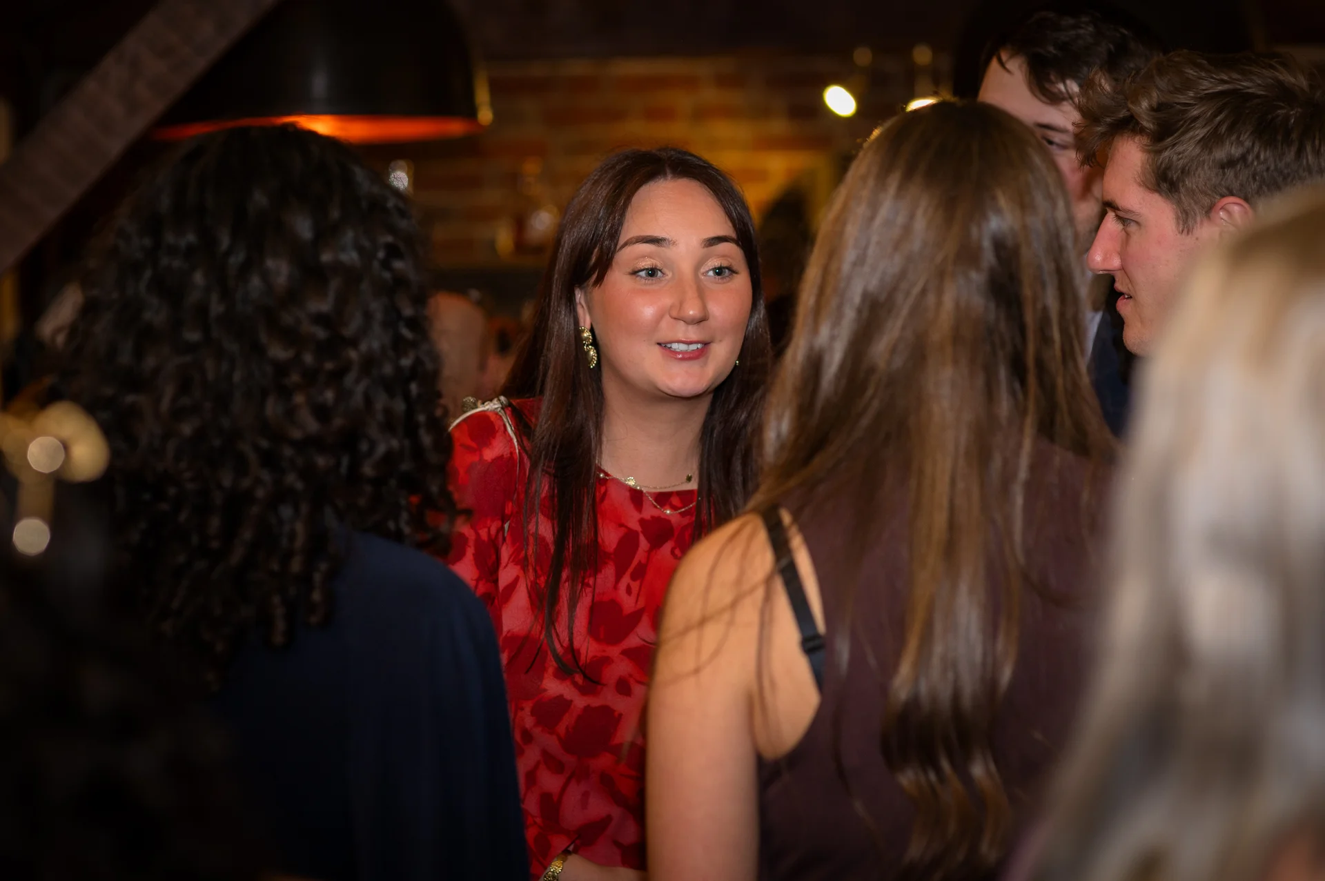 A group of wedding guests converse and mingle together in an indoor venue with an exposed brick wall background.