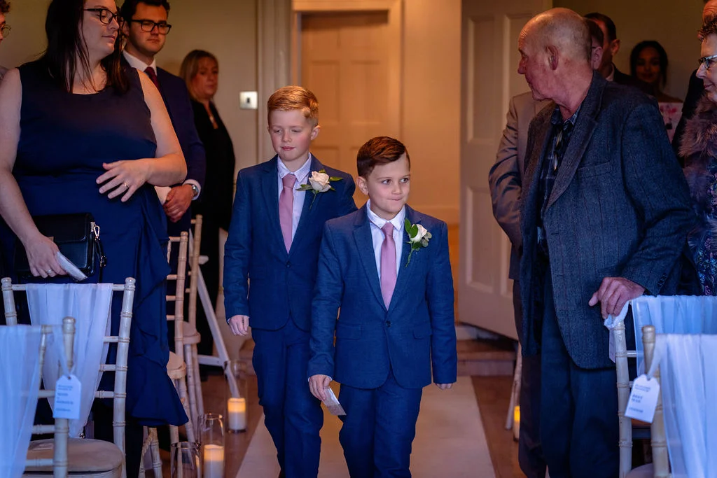 Two young pageboys in matching blue suits and pink ties walk down the aisle past seated wedding guests.