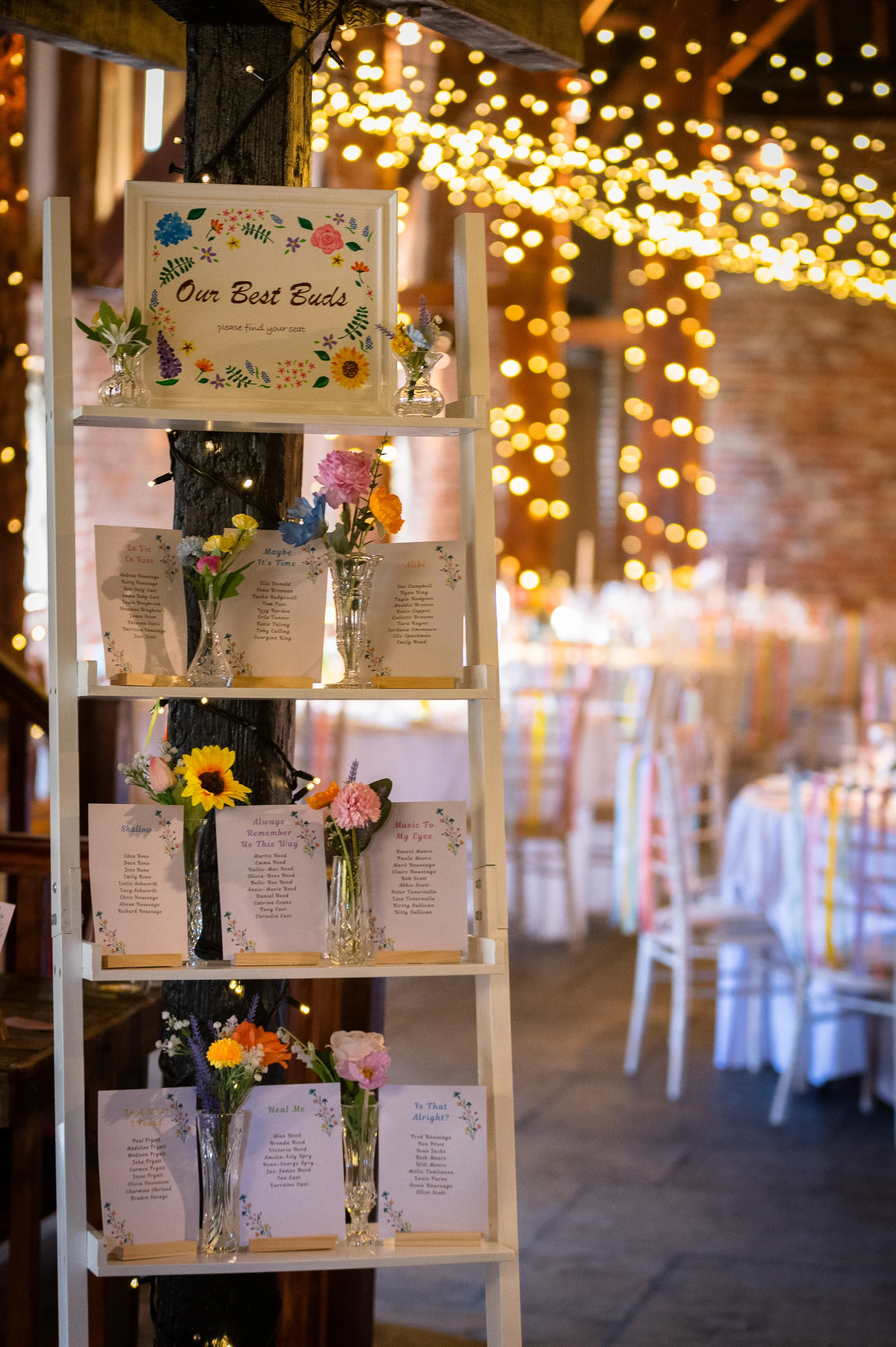 A creative seating plan is displayed on a white wooden ladder adorned with bud vases and floral stationery inside a rustic barn illuminated by a glowing fairy light canopy.