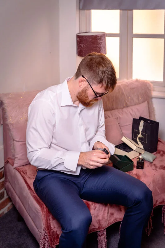 A groom with a ginger beard wearing a white shirt and trousers sits on a pink sofa by a window, carefully adjusting his watch.