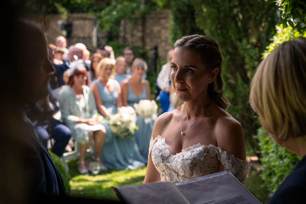 A radiant bride in an off-the-shoulder floral appliqué gown stands smiling during an outdoor celebrant-led ceremony, with guests and blue-clad bridesmaids seated in the sunlit background.