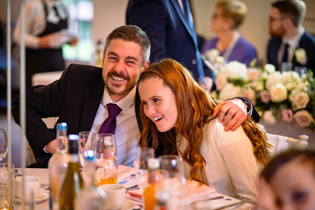 A man in a suit with a purple tie and a woman with long hair laugh together while seated at a wedding reception table.