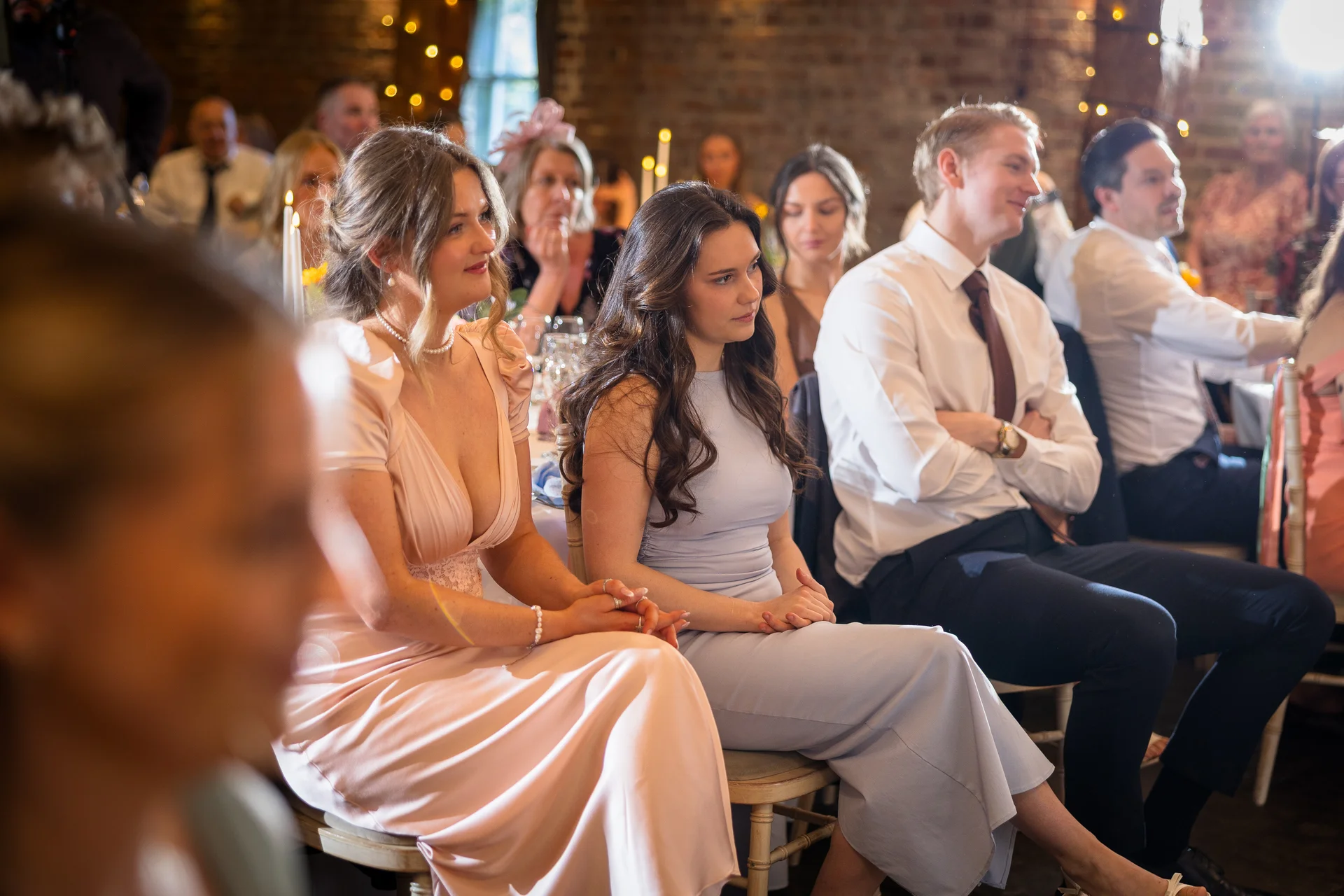A candid shot of wedding guests seated at tables during the reception inside a rustic brick barn venue, watching attentively.
