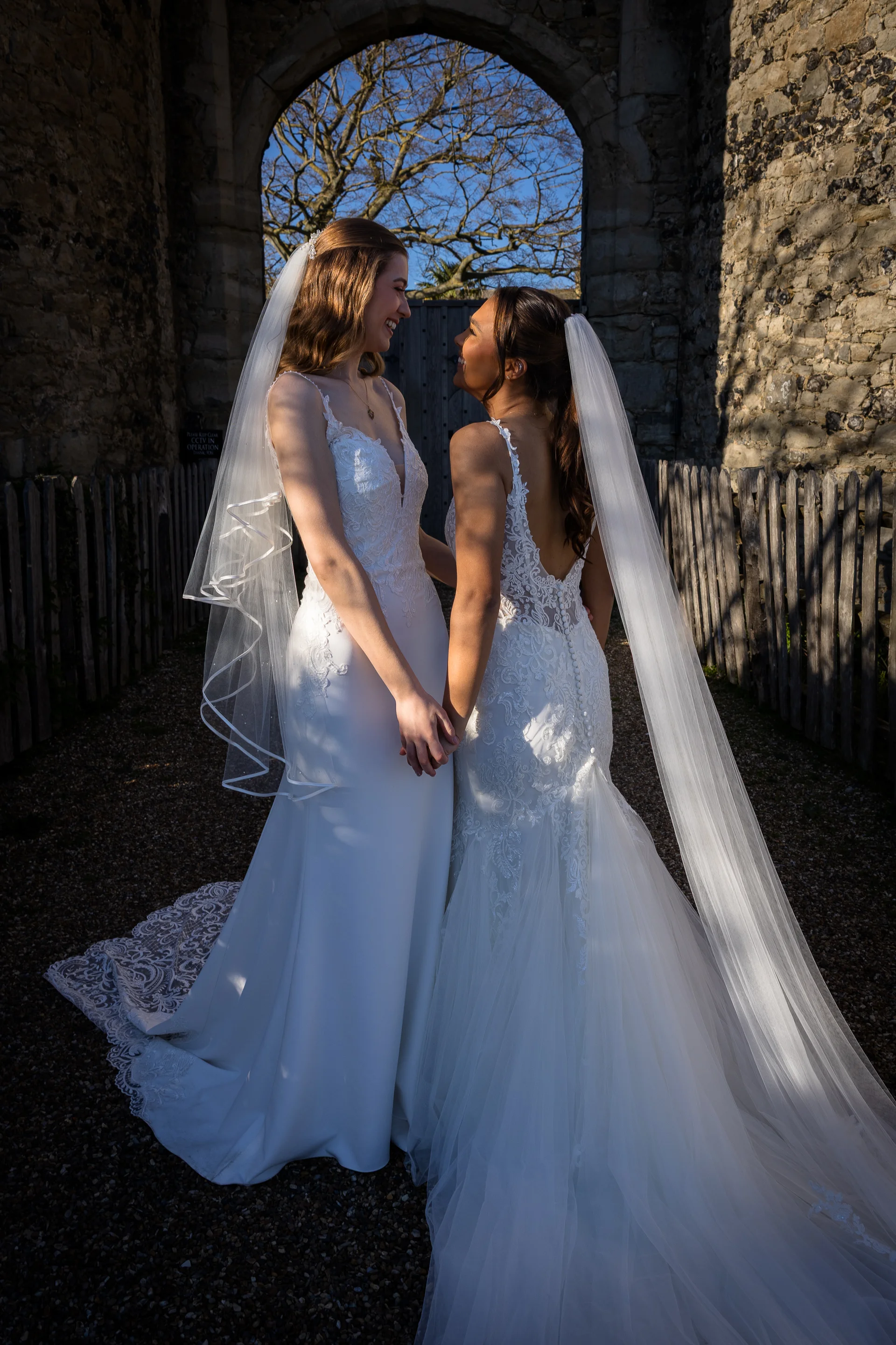 Two smiling brides stand hand in hand gazing at each other beneath a stone castle archway on a sunny wedding day.