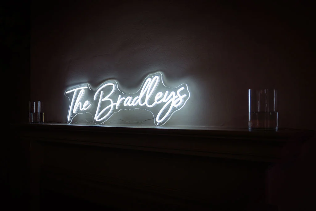 A glowing white custom neon sign reading 'The Bradleys' sits atop a dark fireplace mantelpiece during an evening wedding reception.