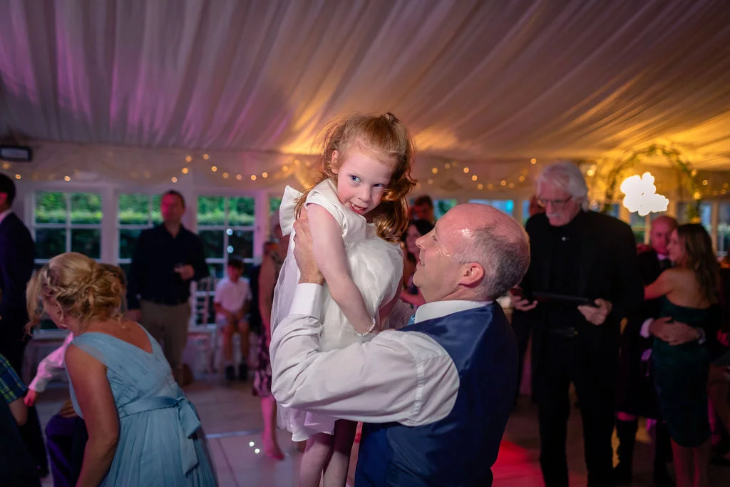 A guest joyfully lifts a young flower girl into the air on a lit marquee dance floor during a wedding reception.