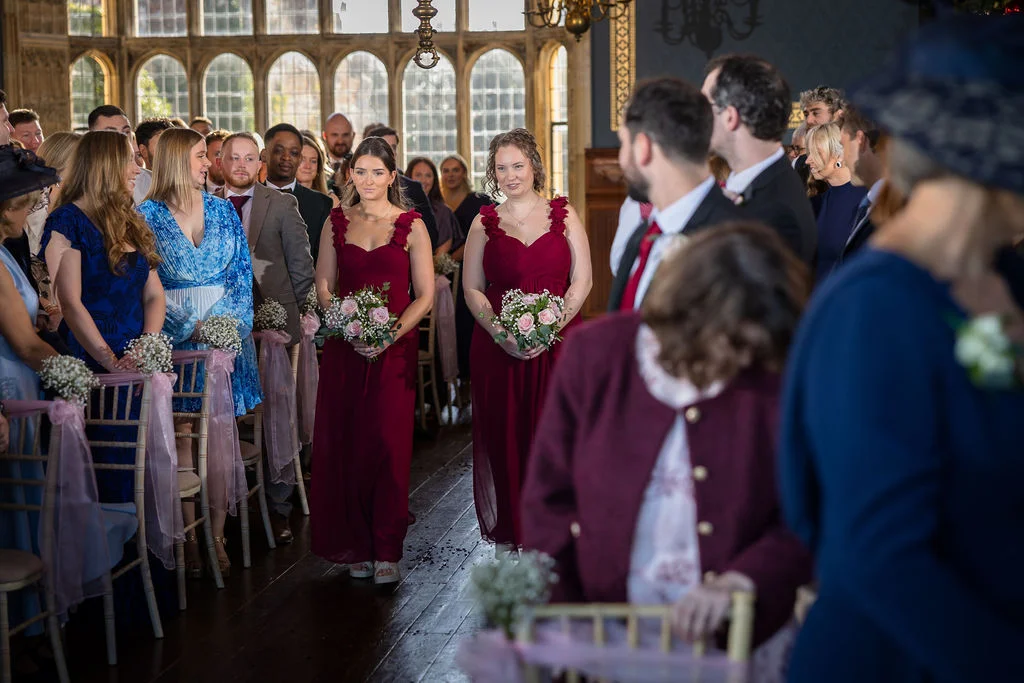 Two bridesmaids in matching burgundy dresses walk down the aisle carrying small bouquets of roses and baby's breath during a country house wedding ceremony.