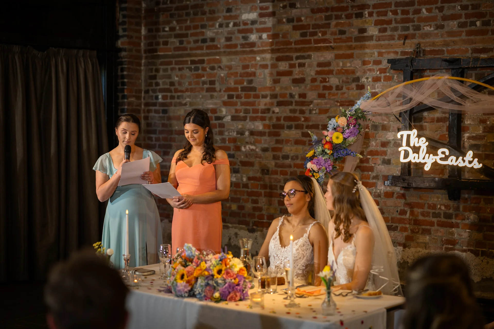 A bridesmaid delivers a speech from notes using a microphone during the wedding reception while two brides look on from the top table in an industrial venue with exposed brick.