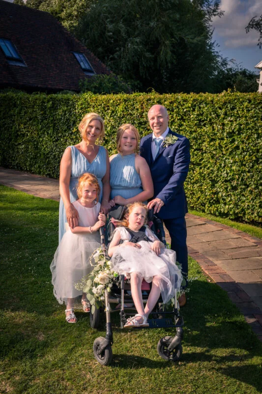 A smiling wedding party including a groom in a navy suit, women in dusty blue dresses, and children posing outdoors on a sunny lawn by a tall green hedge.