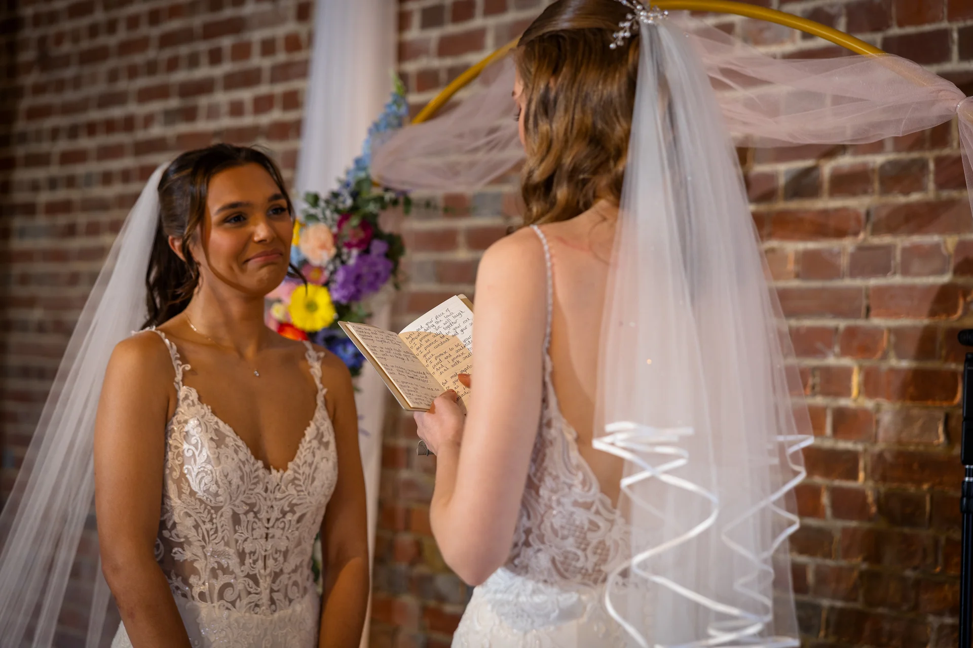 Two brides share a deeply emotional moment during their indoor ceremony as one reads handwritten vows from a small booklet in front of an exposed brick wall.