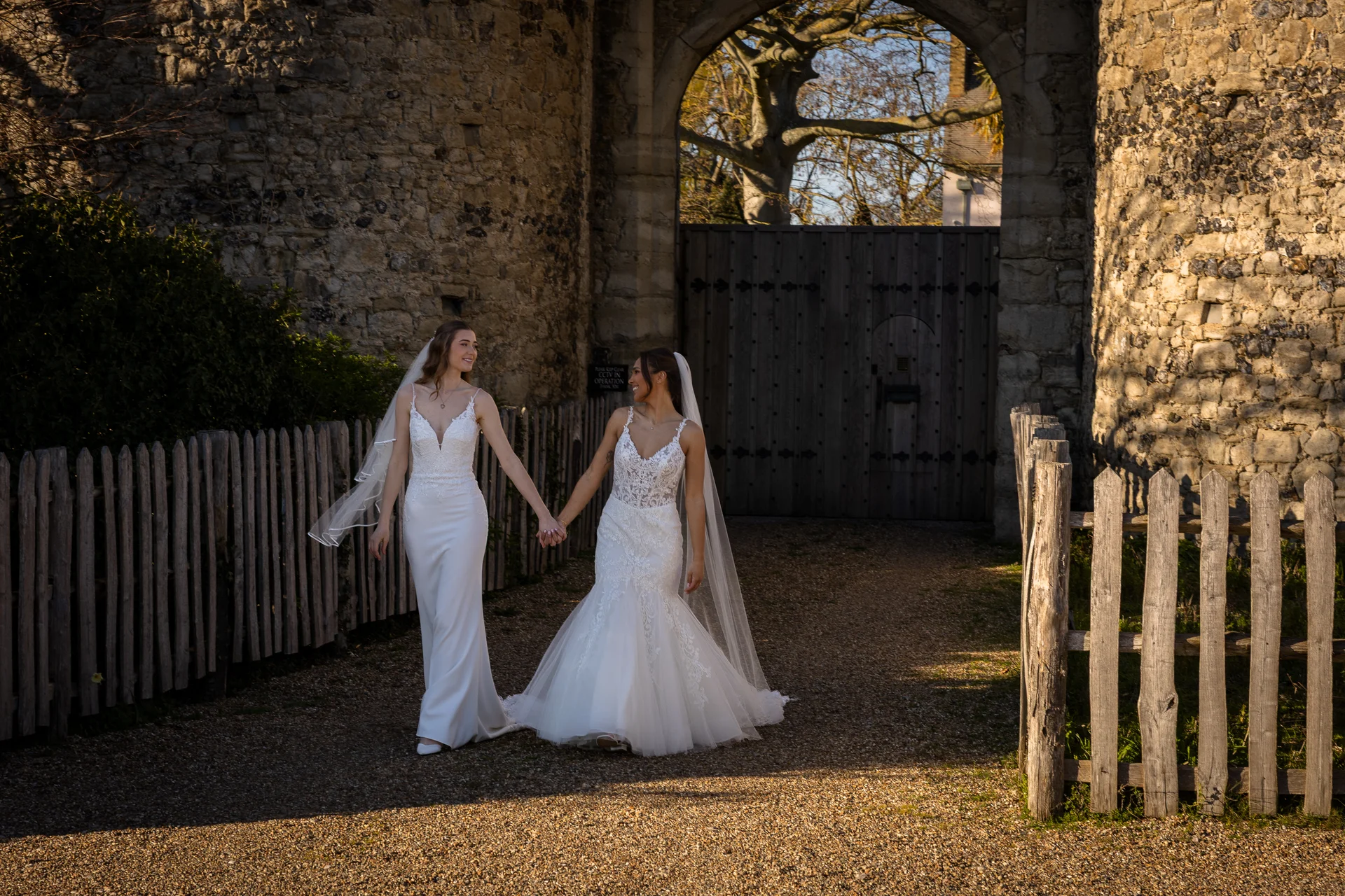 Two brides hold hands and smile at each other while walking down a gravel path outside a historic stone castle gateway during golden hour.