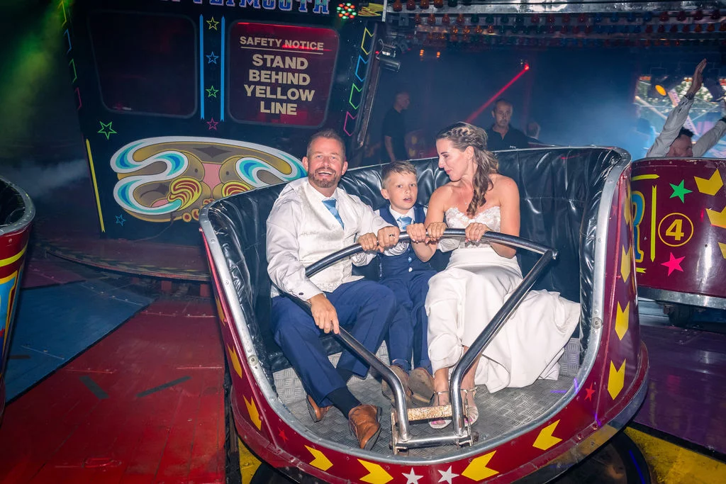 A newlywed couple and a young page boy share a joyful ride on a bright funfair fairground attraction during their evening wedding reception.