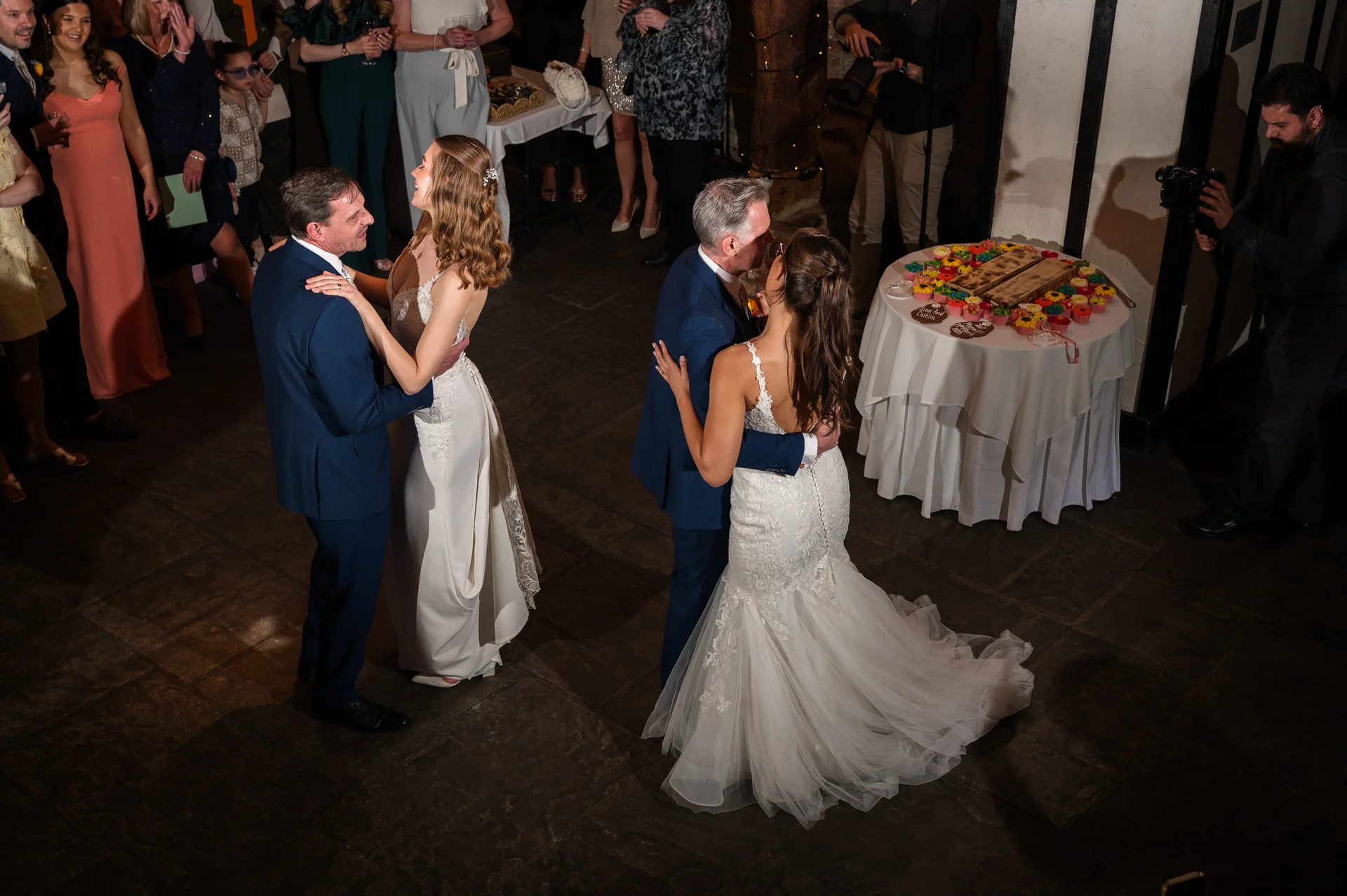 Two couples share their first dance on a stone floor during a wedding reception, surrounded by watching guests and a small table of cupcakes.