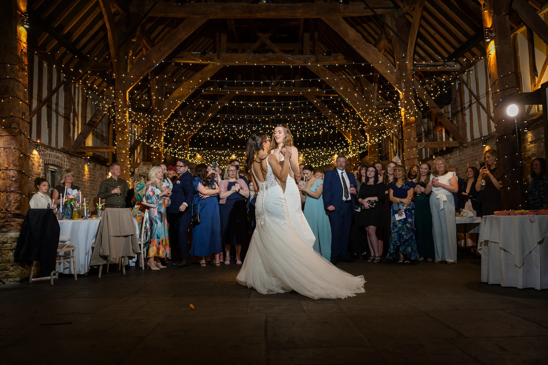Two brides share their first dance in the centre of a large rustic oak-framed barn illuminated by a canopy of fairy lights as wedding guests watch from the sidelines.