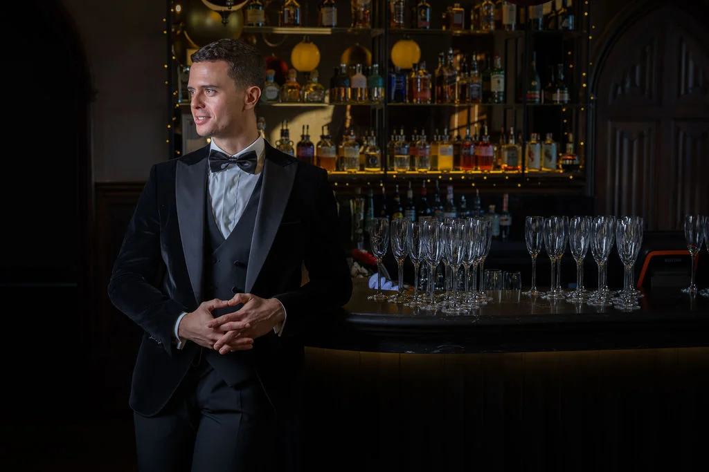 A stylish groom in a black velvet tuxedo and bow tie leans against a dimly lit bar counter filled with champagne flutes.