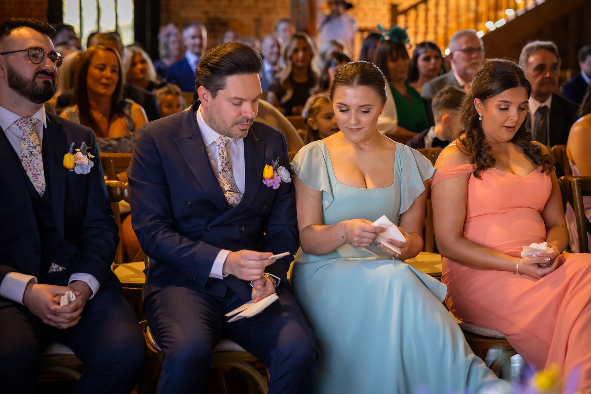 A group of wedding guests sitting together in a venue, looking down thoughtfully while holding small cards or tissues during the ceremony.