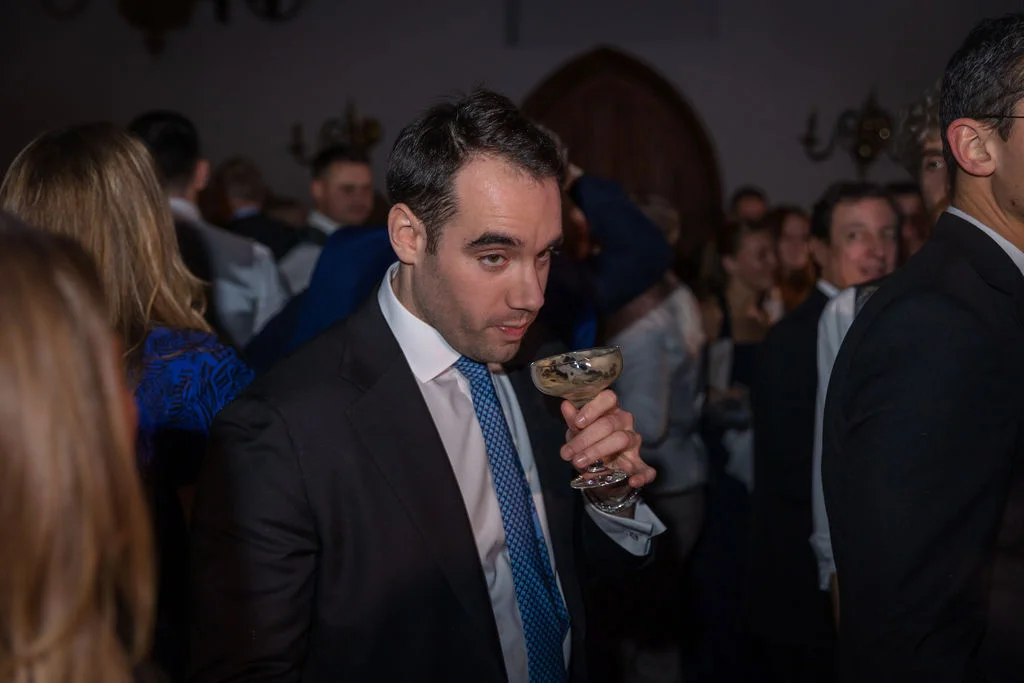 A guest in a black suit and patterned tie inspects a champagne coupe amidst a crowded evening reception.