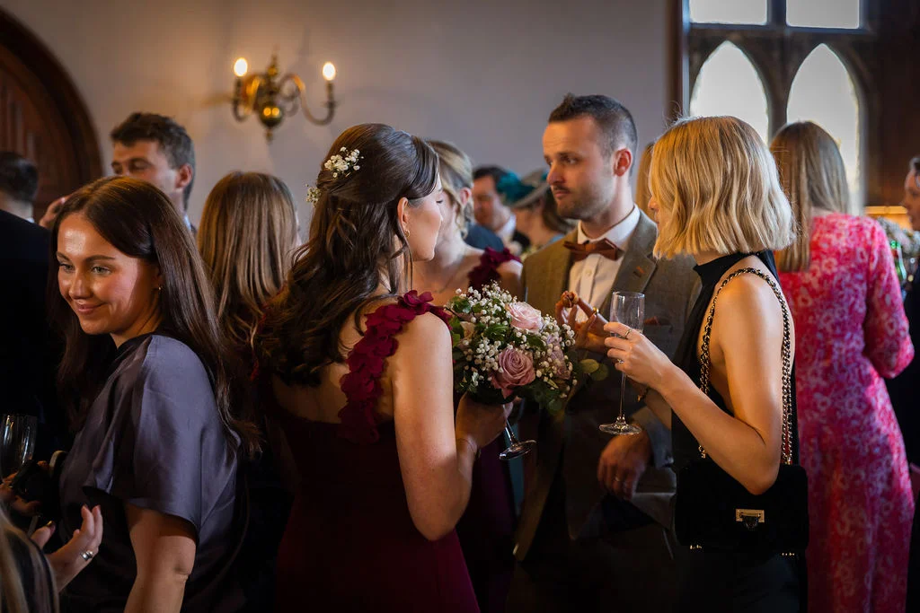 Wedding guests mingle and chat during a drinks reception inside a country house venue, holding champagne flutes and bouquets.