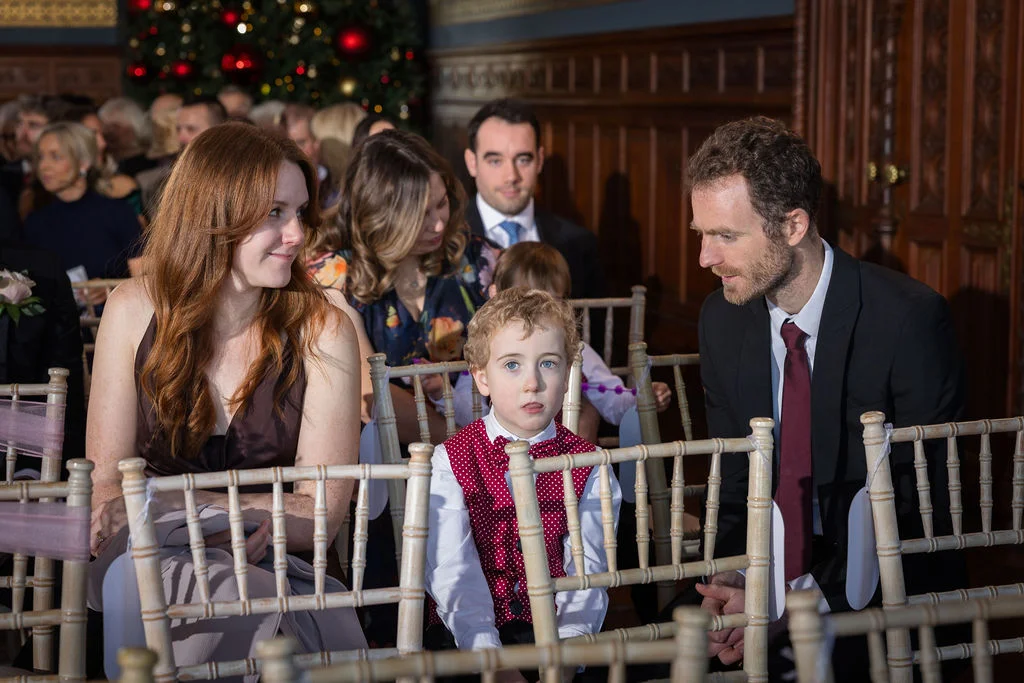 Family members and a young page boy in a red patterned waistcoat are seated together on wooden chairs inside a wood-panelled venue during a winter wedding.