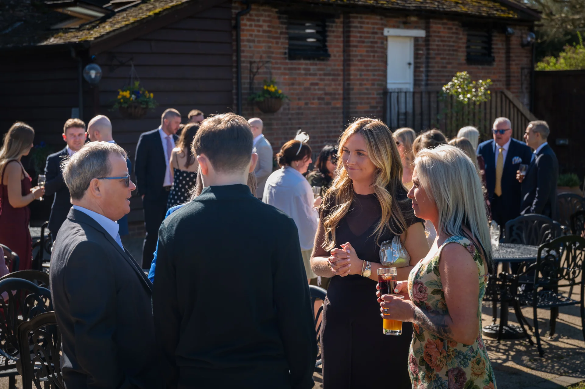 Wedding guests mingle and converse in a sunlit brick courtyard outside a rustic barn venue during the drinks reception.