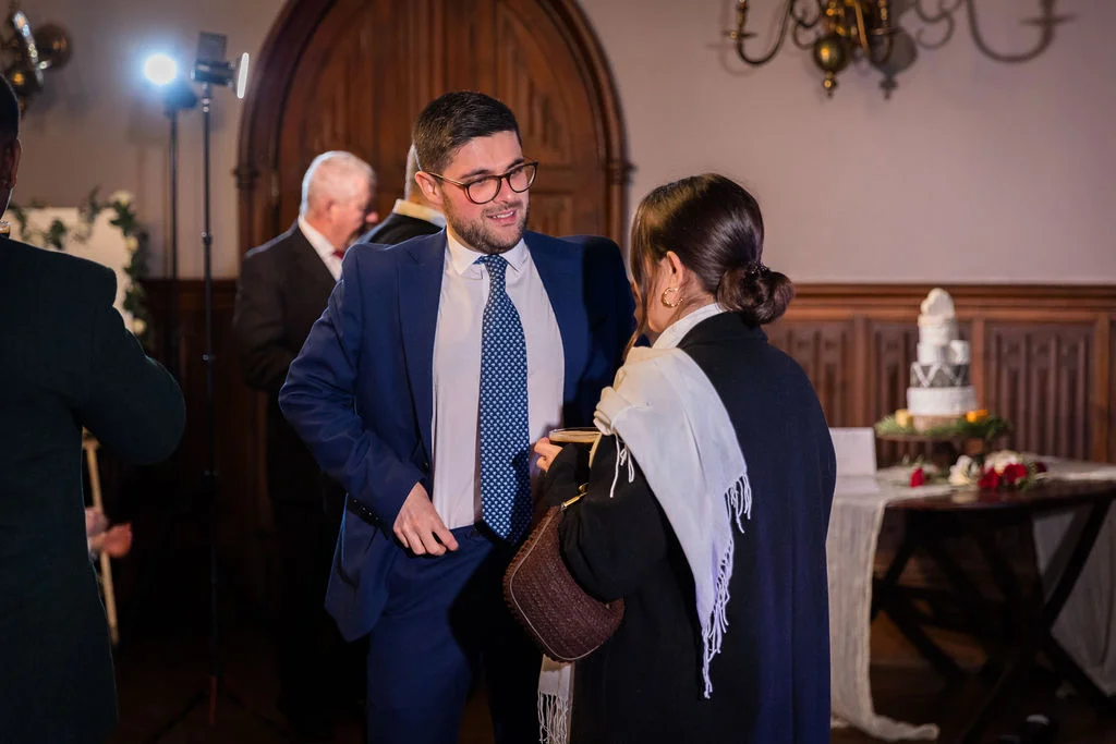 A guest in a sharp navy suit with a patterned tie smiles while talking to another guest holding a drink and handbag during a wedding reception in a wood-panelled room.