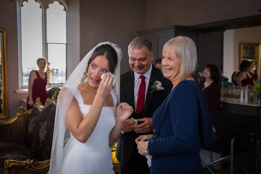 An emotional bride gently wipes a tear away with a tissue while standing alongside her smiling father and mother inside a historic room with tall arched windows.