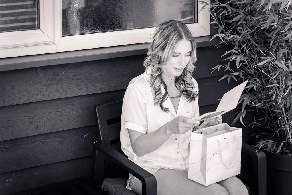 A smiling bride reads a heartfelt letter while sitting outdoors on a chair with a gift bag during morning preparations.