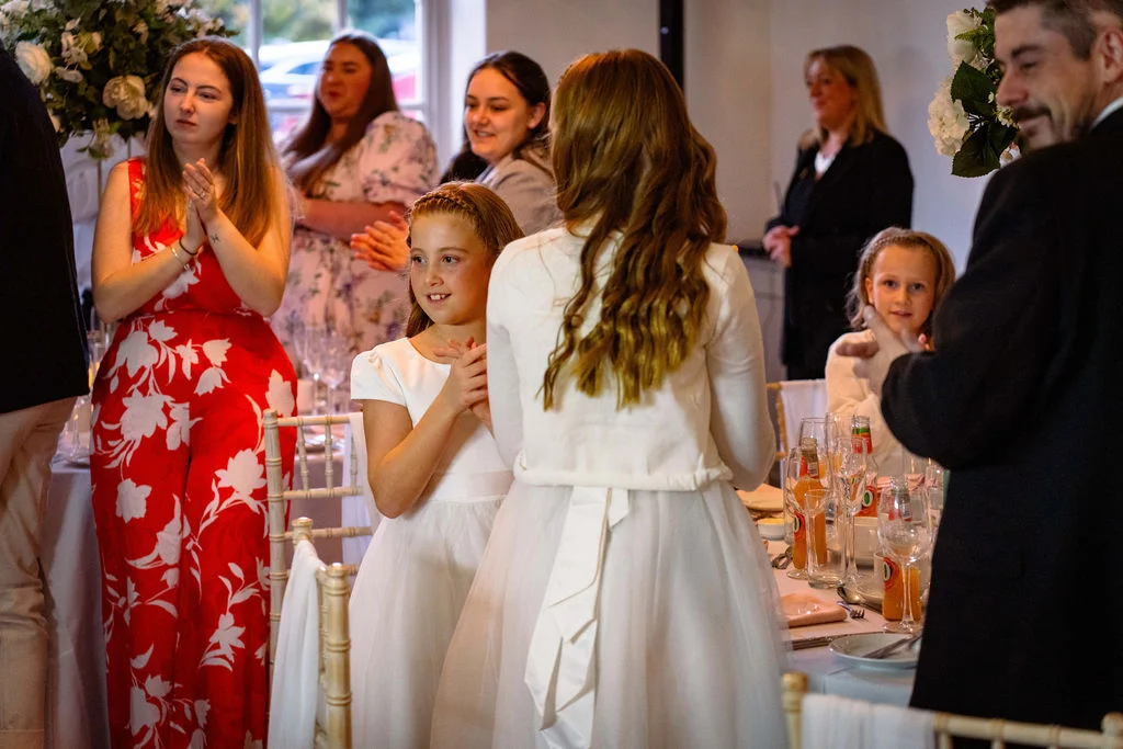 Guests and young flower girls smile and applaud during wedding speeches at a brightly lit indoor reception.