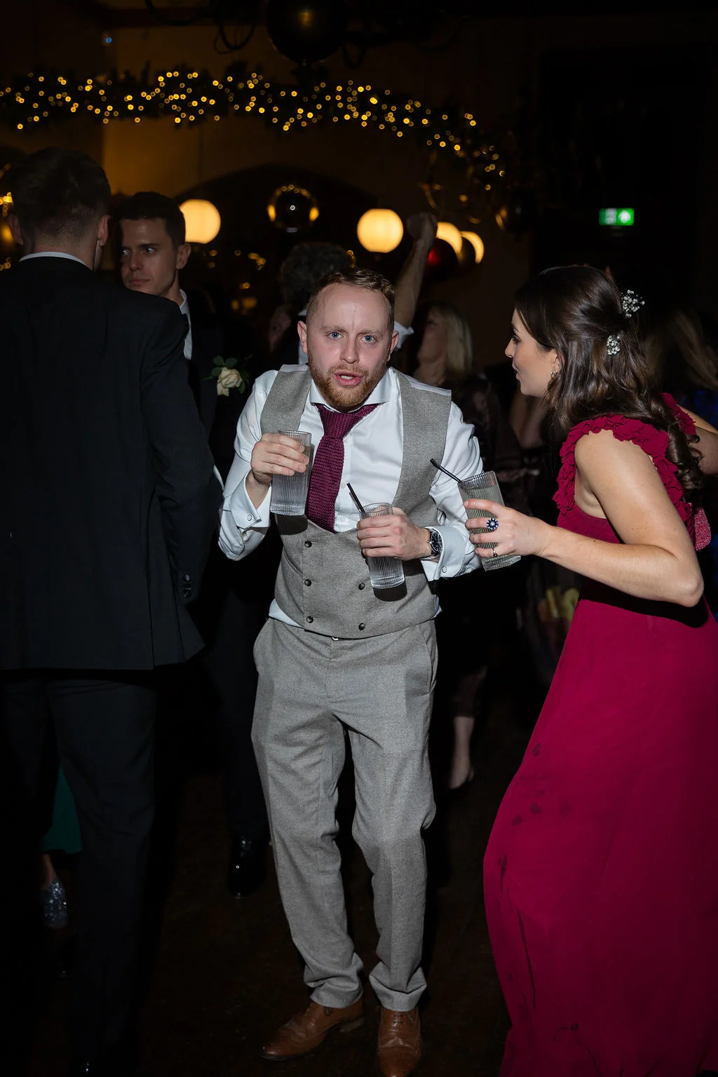 A groom in a tweed waistcoat and burgundy tie holds two drinks while dancing alongside guests at an evening wedding reception lit by fairy lights.