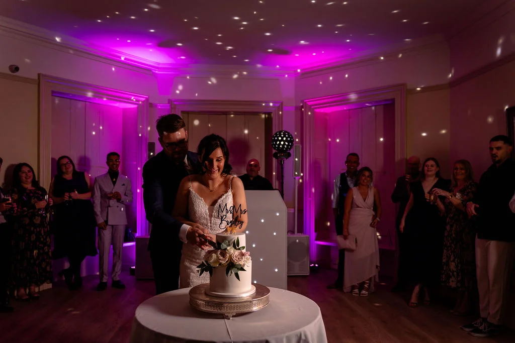 The bride and groom cut their two-tier wedding cake together while surrounded by guests in a reception room bathed in vibrant pink lighting and disco ball reflections.