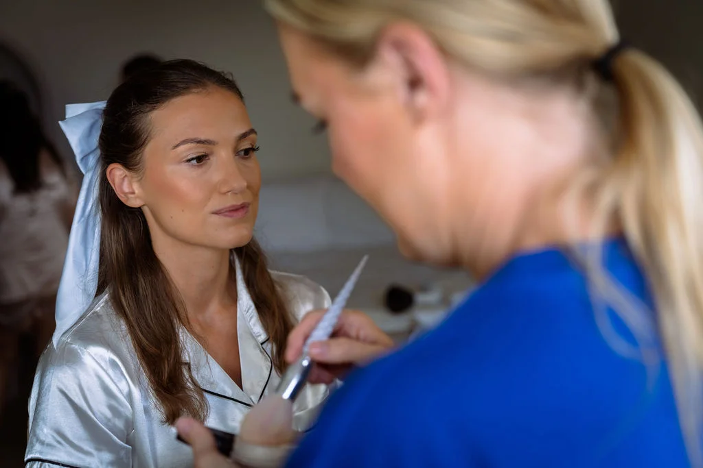 A makeup artist applies finishing touches to a bride wearing satin pyjamas and a bow in her hair during morning bridal preparations.