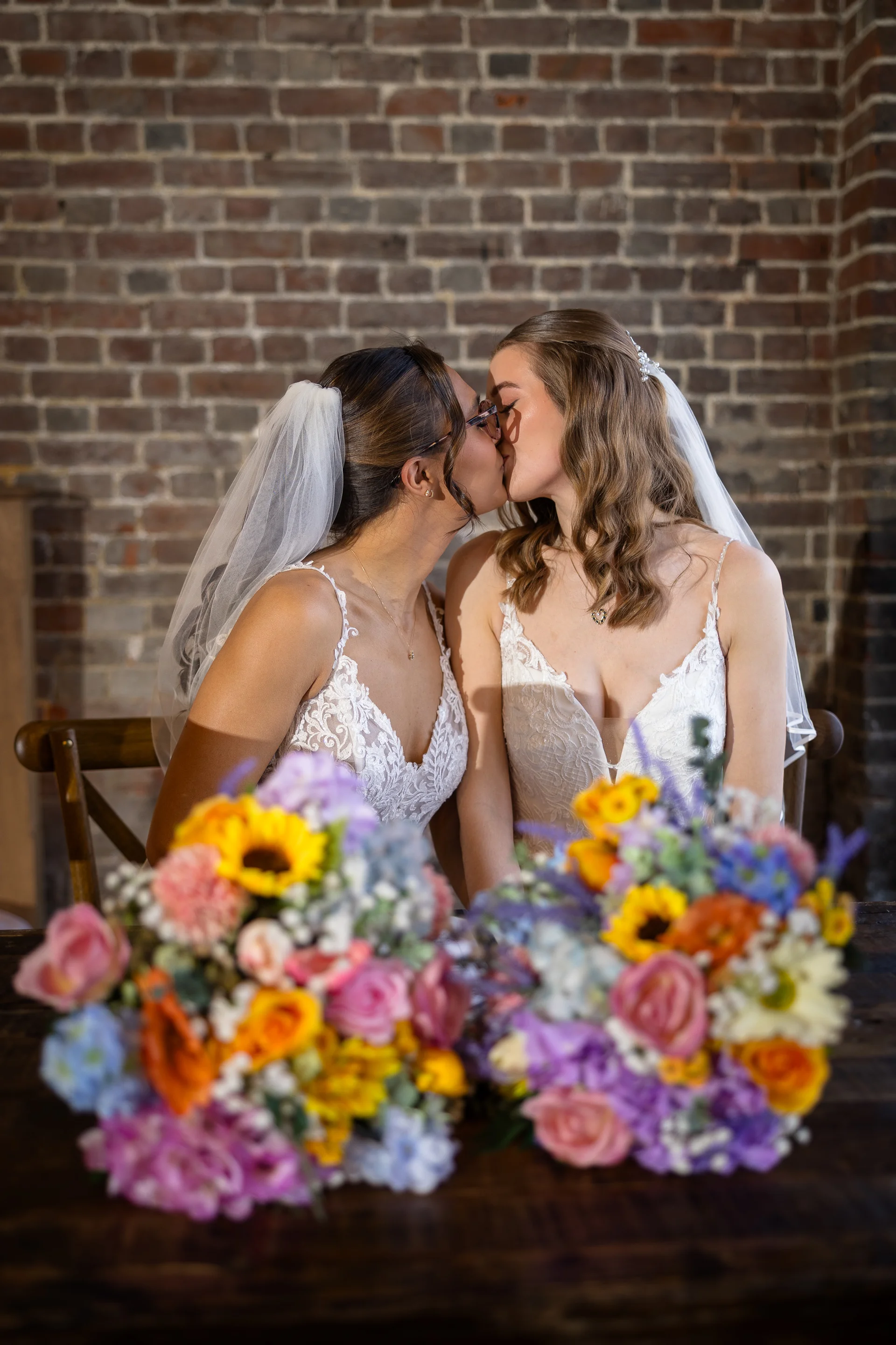 Two smiling brides share a tender kiss in front of an exposed brick wall, framed by colourful wildflower bouquets on a rustic wooden table.