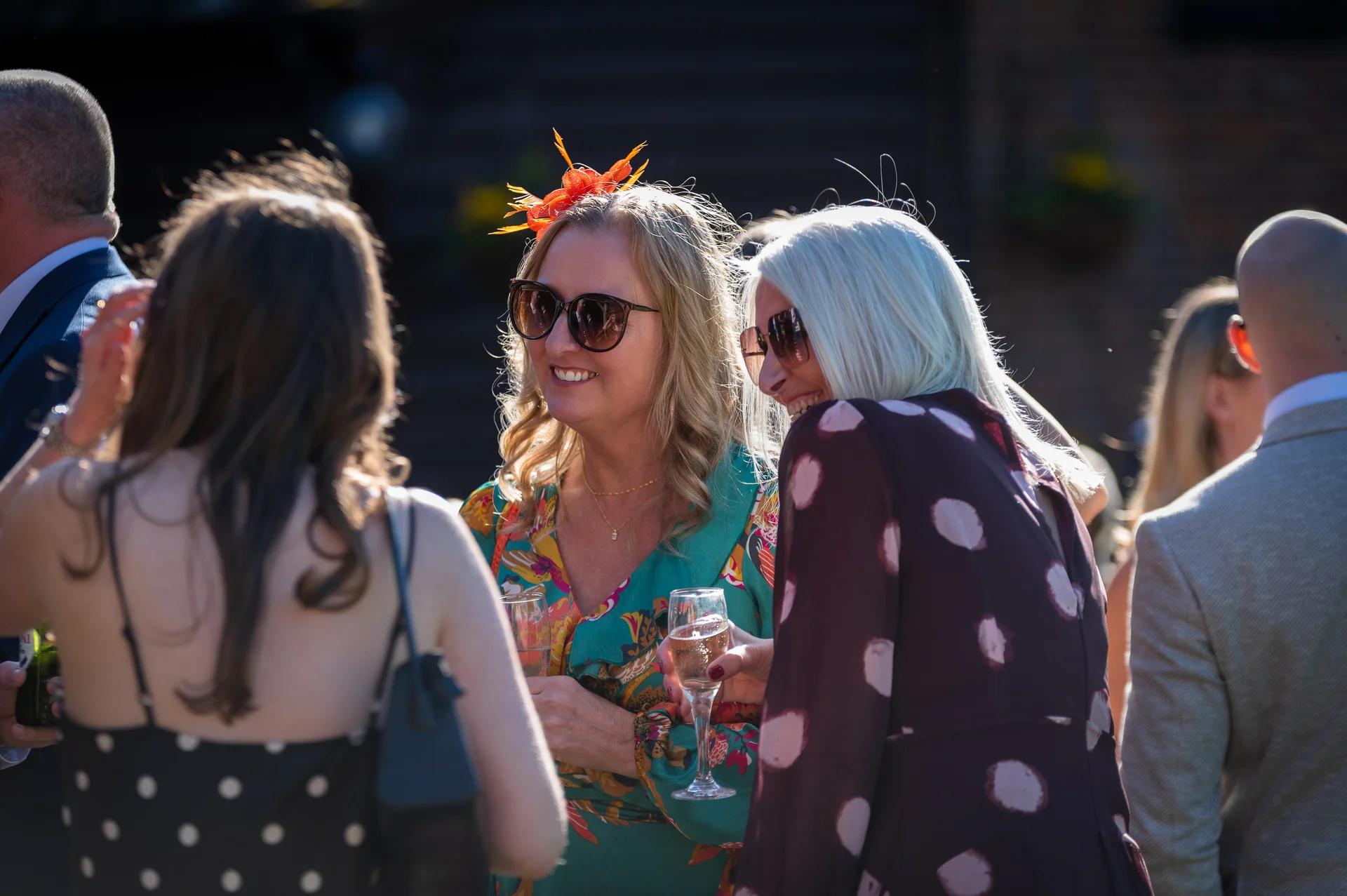 Wedding guests wearing stylish outfits and sunglasses mingle outdoors in bright sunlight while holding glasses of champagne during the drinks reception.