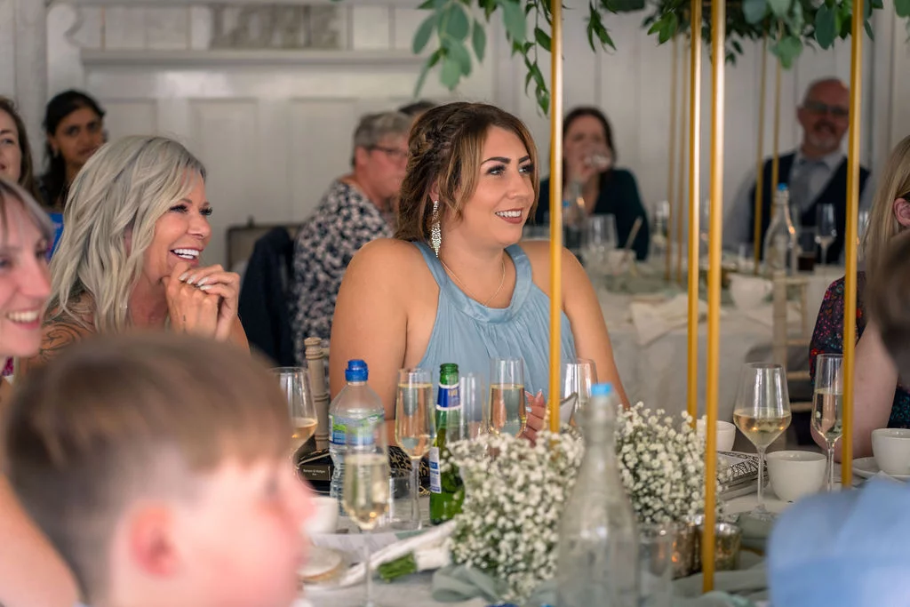 Wedding guests laugh and smile while listening to speeches during the wedding breakfast, seated at a beautifully decorated table with floral arrangements and glassware.