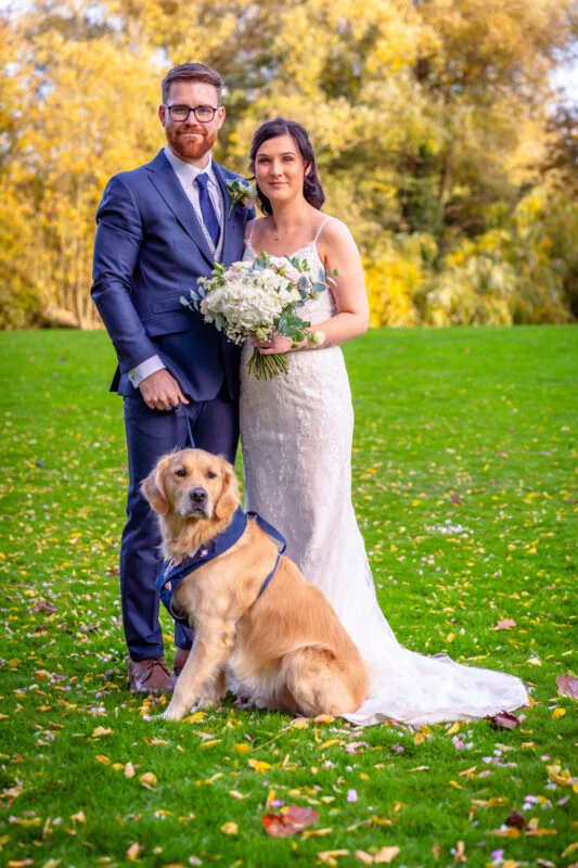 A bride and groom pose on a vibrant green lawn with their golden retriever dog during an autumn wedding portrait session.
